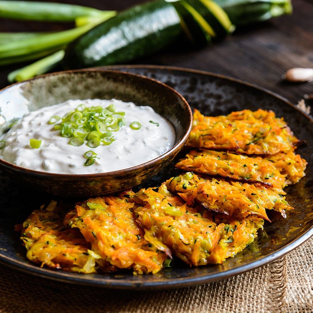 Gebratene Zucchinipuffer mit Dip. Puffer orangebraun, Dip in Schale. Grüne Zwiebeln als Garnitur.