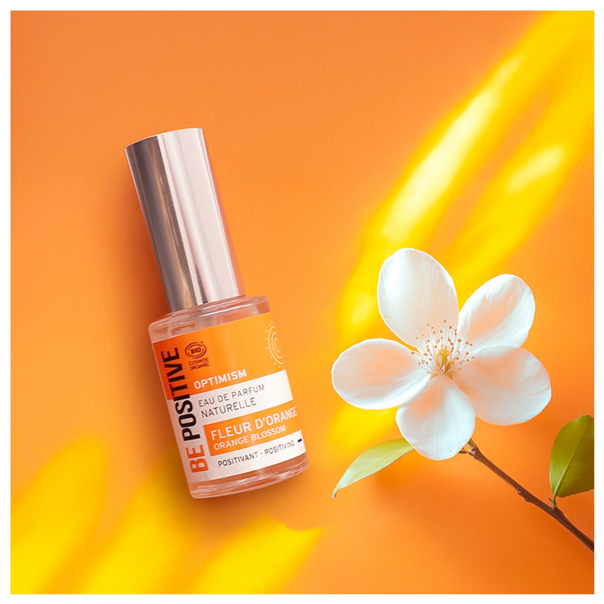 Acorelle Be Positive Eau de Parfum Optimism Fleur d'Oranger neben einer Blüte auf orangefarbenem Hintergrund.