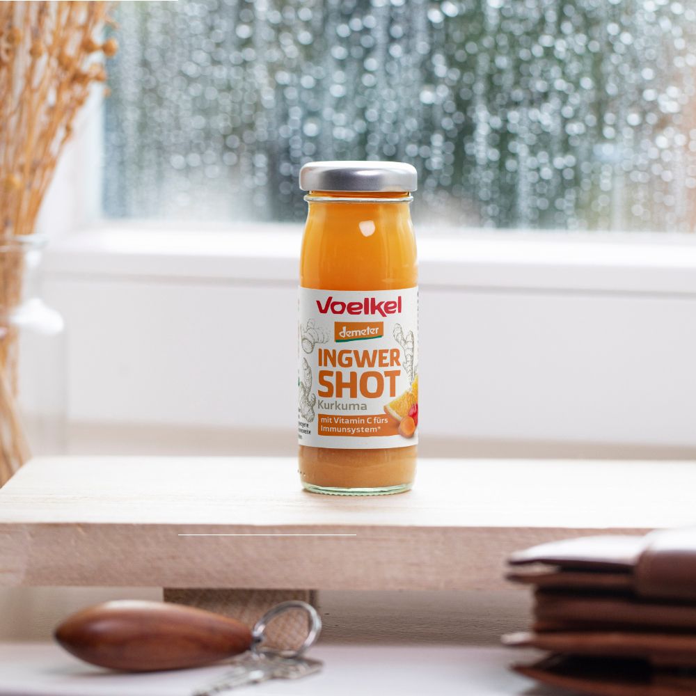 Glasflasche mit gelbem Inhalt. Etikett mit Produktname. Auf Holzbrett vor Fenster. Demeter-Siegel.