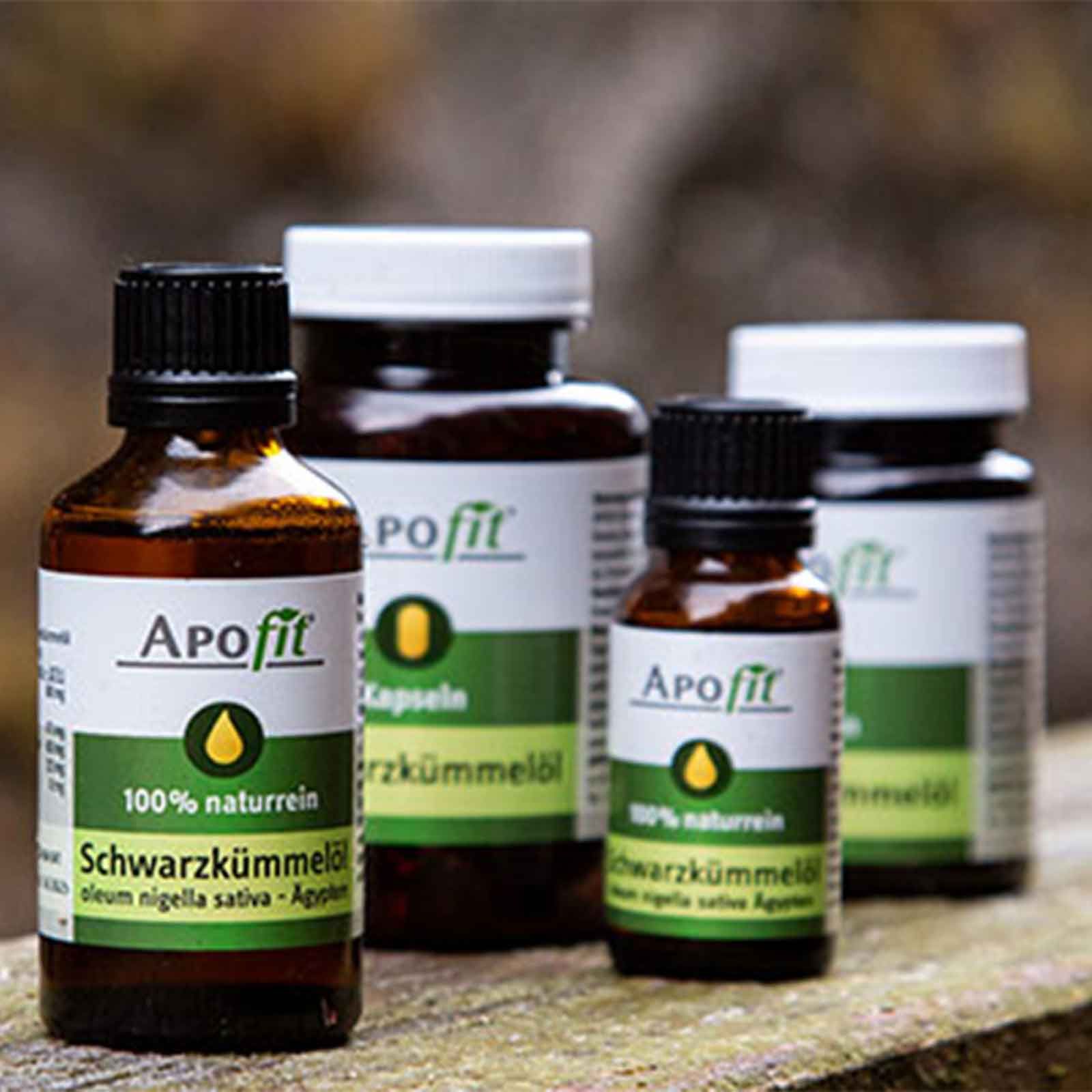 Mehrere APOfit-Produkte, darunter Schwarzkümmelöl-Fläschchen und Kapseln, stehen auf einer Holzoberfläche. Die Flaschen sind braun mit schwarzen Deckeln und weißen Etiketten. Auf den Etiketten sind der Markenname, grüne Bereiche mit gelben Tropfen und Produktbezeichnungen zu sehen. Die Kapseln sind in einer weißen Dose.