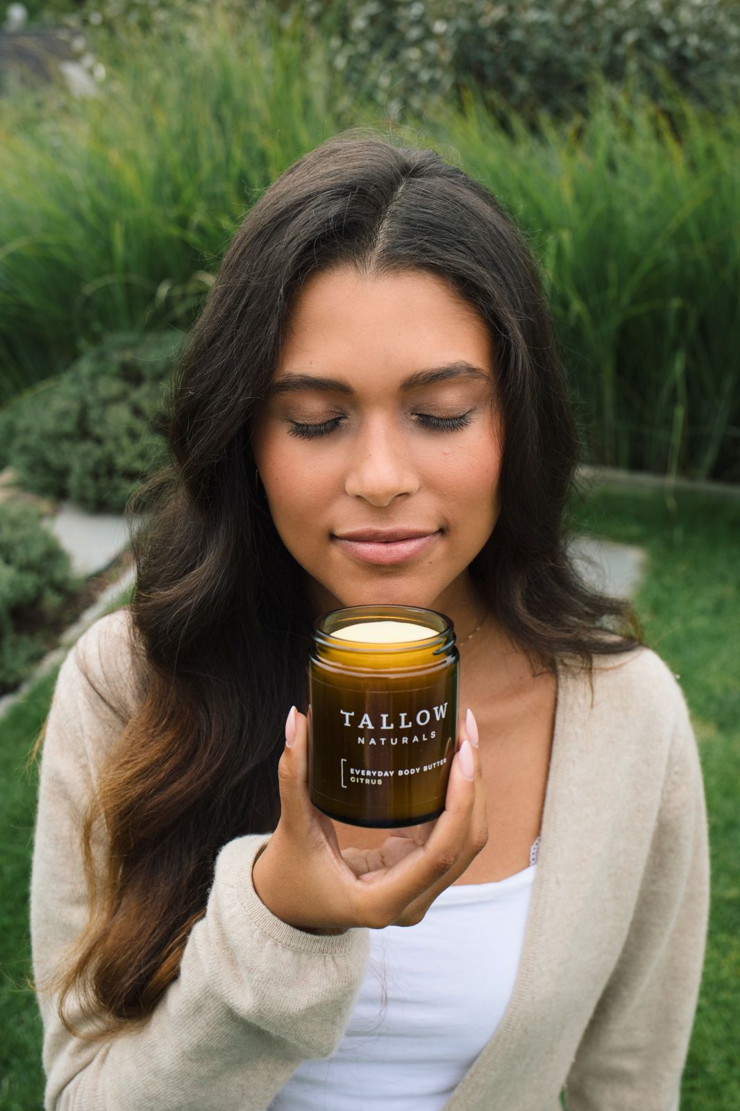 Frau hält eine braune Dose mit schwarzem Deckel. Aufschrift: TALLOW NATURALS, EVERYDAY BODY BUTTER CITRUS. Frau schließt die Augen.