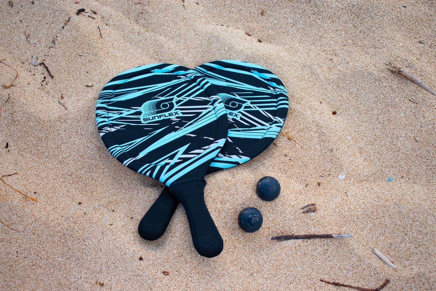 Zwei Schläger und zwei Bälle auf Sand. Die Schläger haben ein blau-schwarzes Muster und das Logo SUNFLEX.