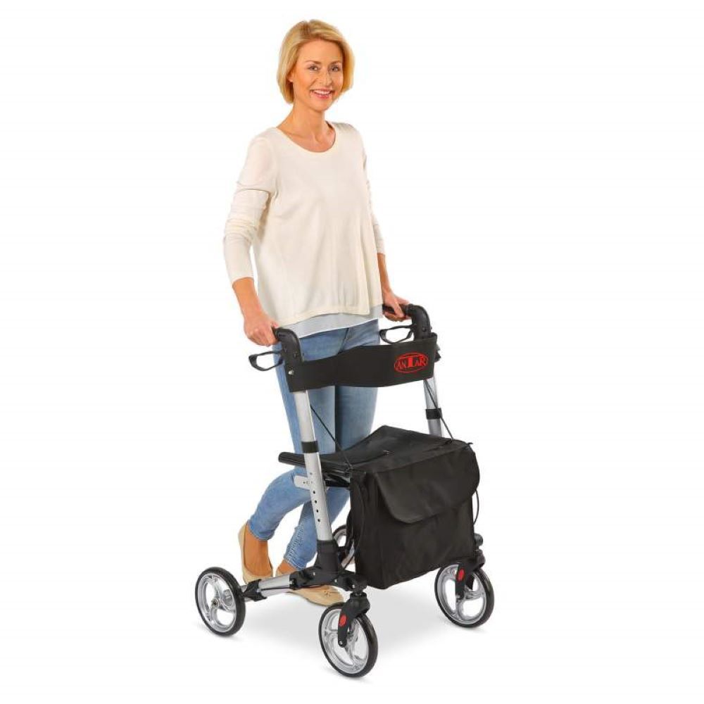 Frau mit Rollator. Silberfarbenes Gestell, schwarze Sitzfläche und Tasche. Drei Räder.