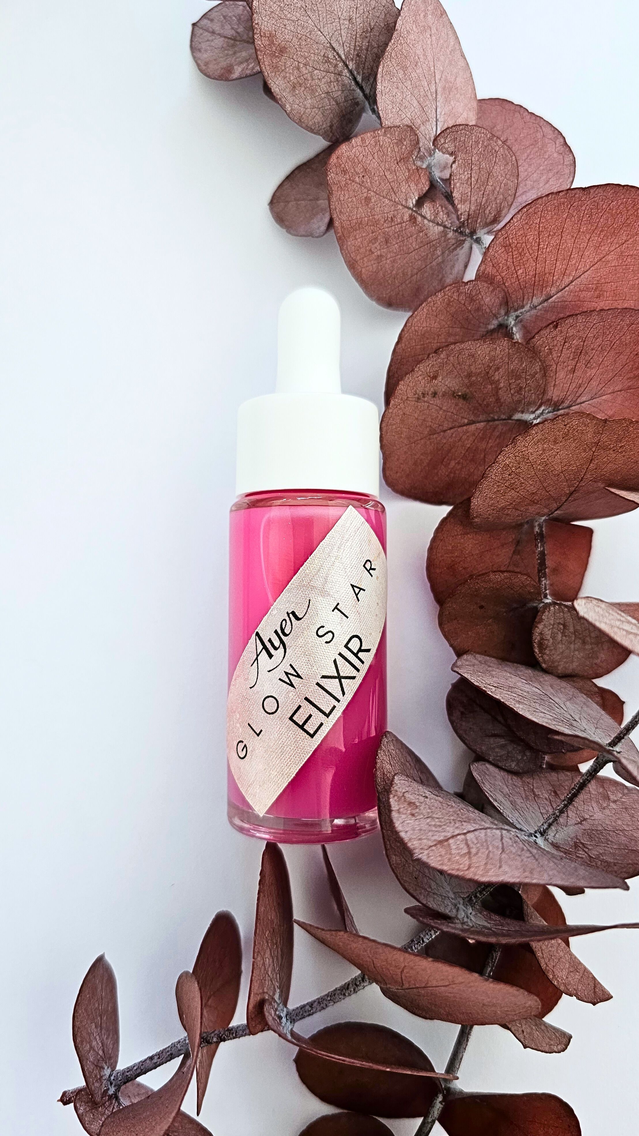 Rosa Flüssigkeit in Glasflasche mit weißem Tropfer, umgeben von getrockneten Blättern. Etikett: Glow Star Elixir.