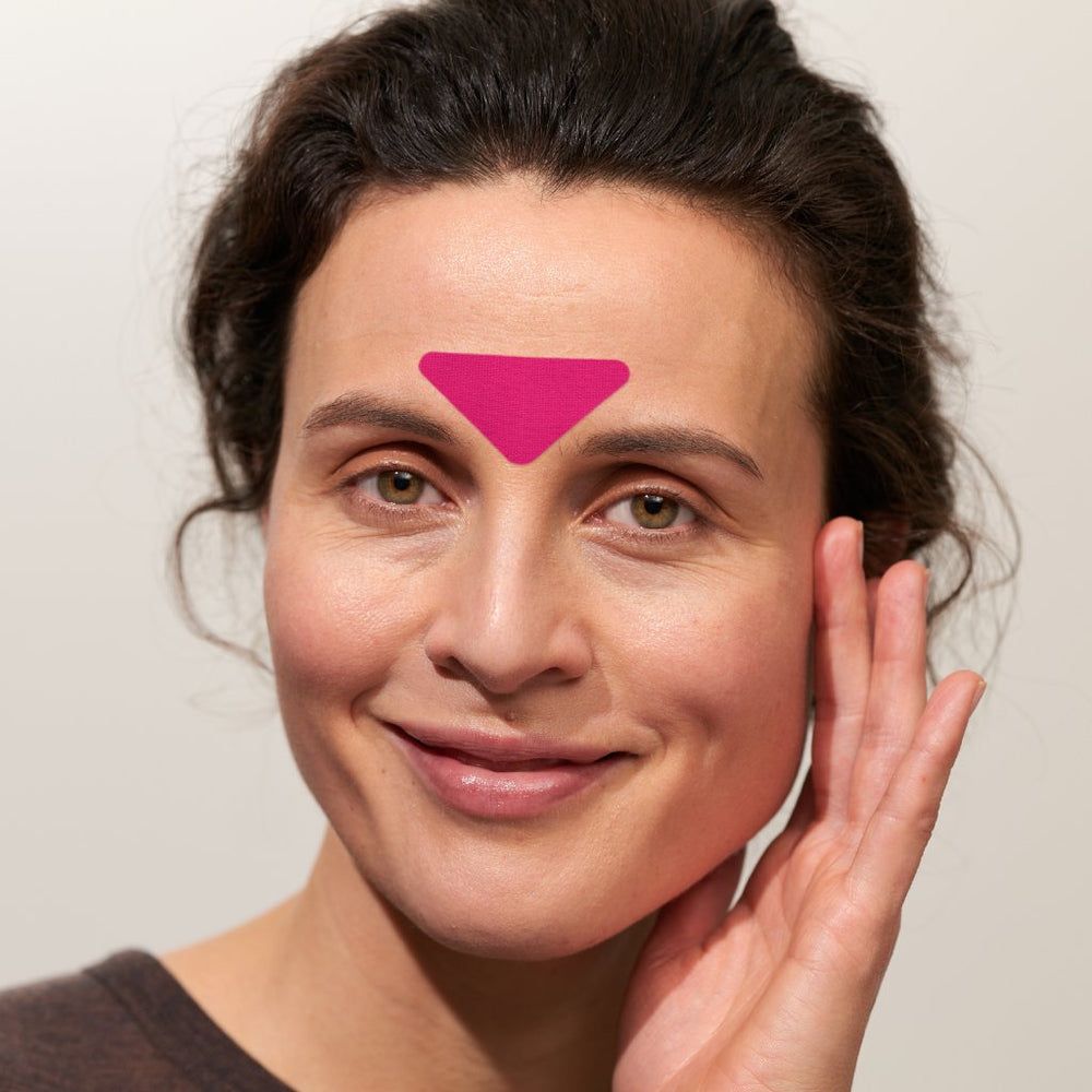 Nahaufnahme einer Frau mit einem rosa Stirn-Gesichtspad. Das Pad ist dreieckig. Die Frau lächelt.