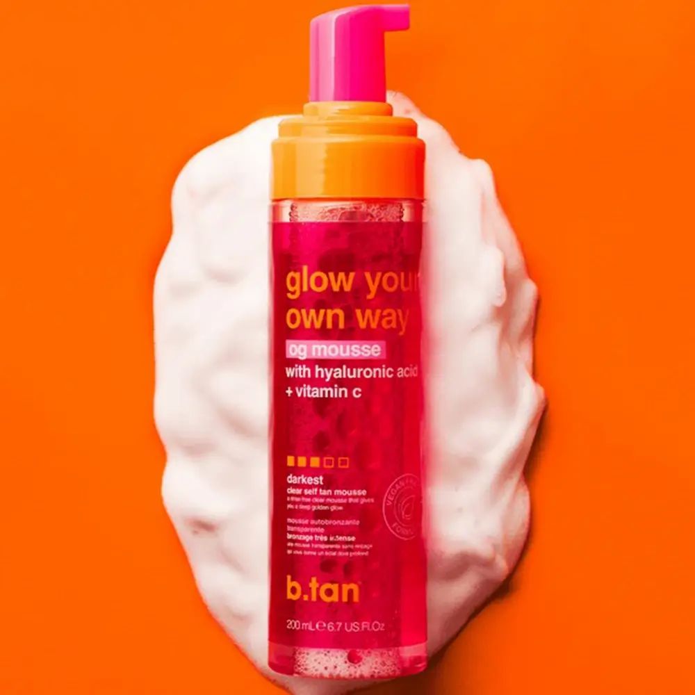 Rosa Flasche mit orangefarbenem Deckel auf weißem Schaum. Aufschrift: glow your own way, og mousse, mit Hyaluronsäure + Vitamin C. Marke: b.tan.