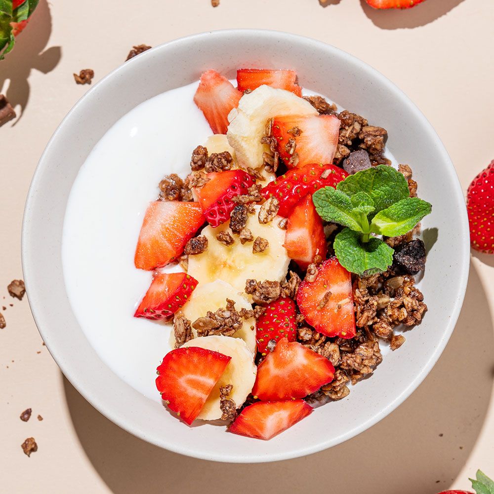 Weiße Schüssel mit Joghurt, Granola, Erdbeeren und Bananenscheiben. Mit Minze garniert. Schatten.