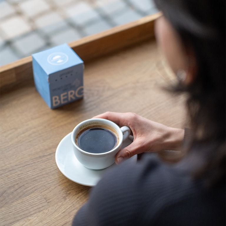 Person trinkt Kaffee aus weißer Tasse mit Untertasse. Blaue BERGBRAND-Box im Hintergrund.