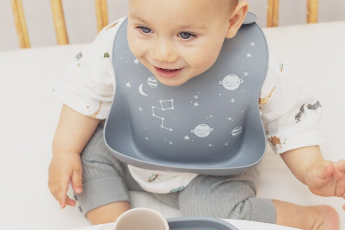 Baby trägt graues Silikon-Lätzchen mit Sternen-Motiv. Das Kind sitzt vor einem Tisch. Sterne, Mond und Planeten sind abgebildet.