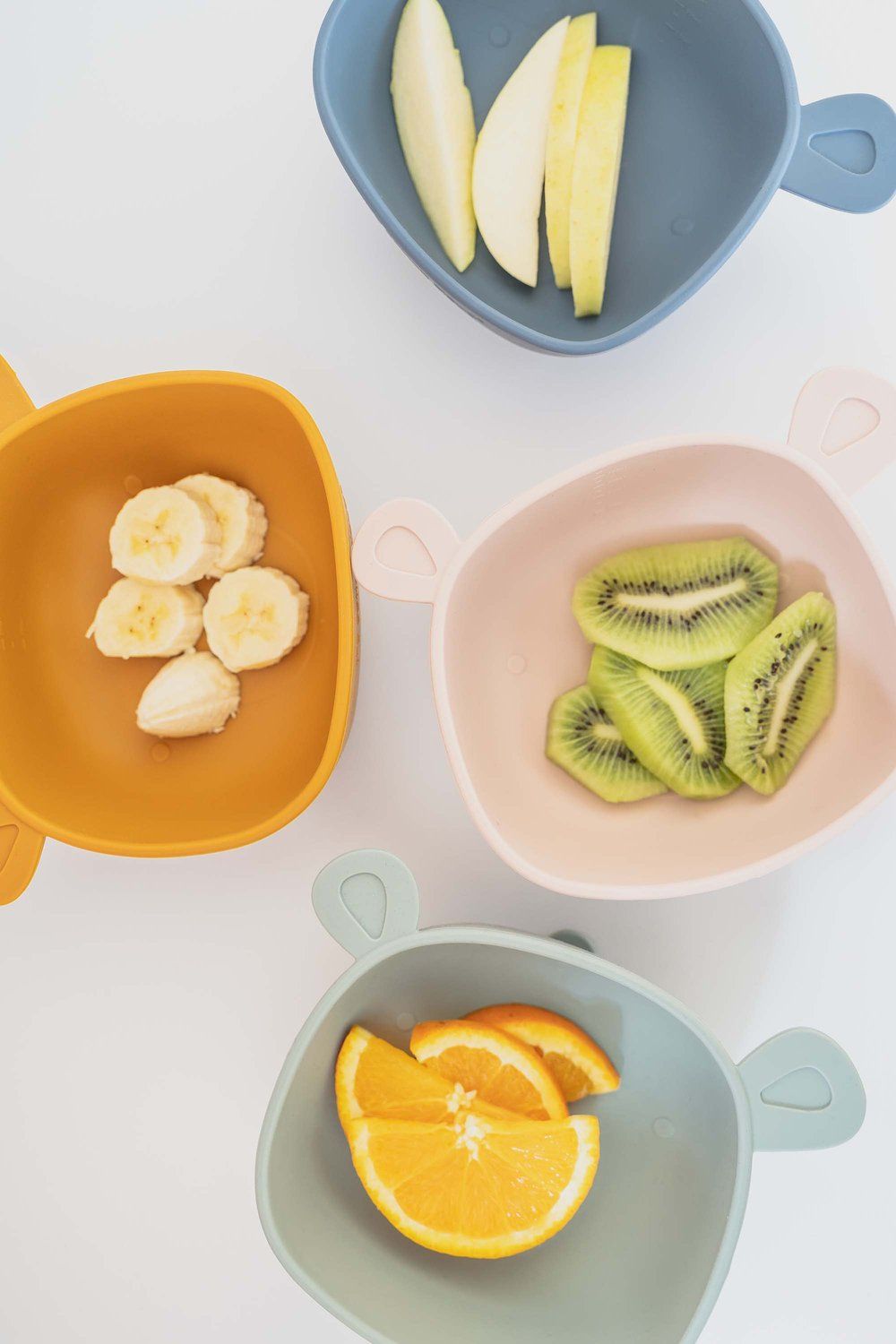Vier Schüsseln mit Ohren, gefüllt mit Obst. Farben: Gelb, Rosa, Blau und Grün. Auf weißem Hintergrund.