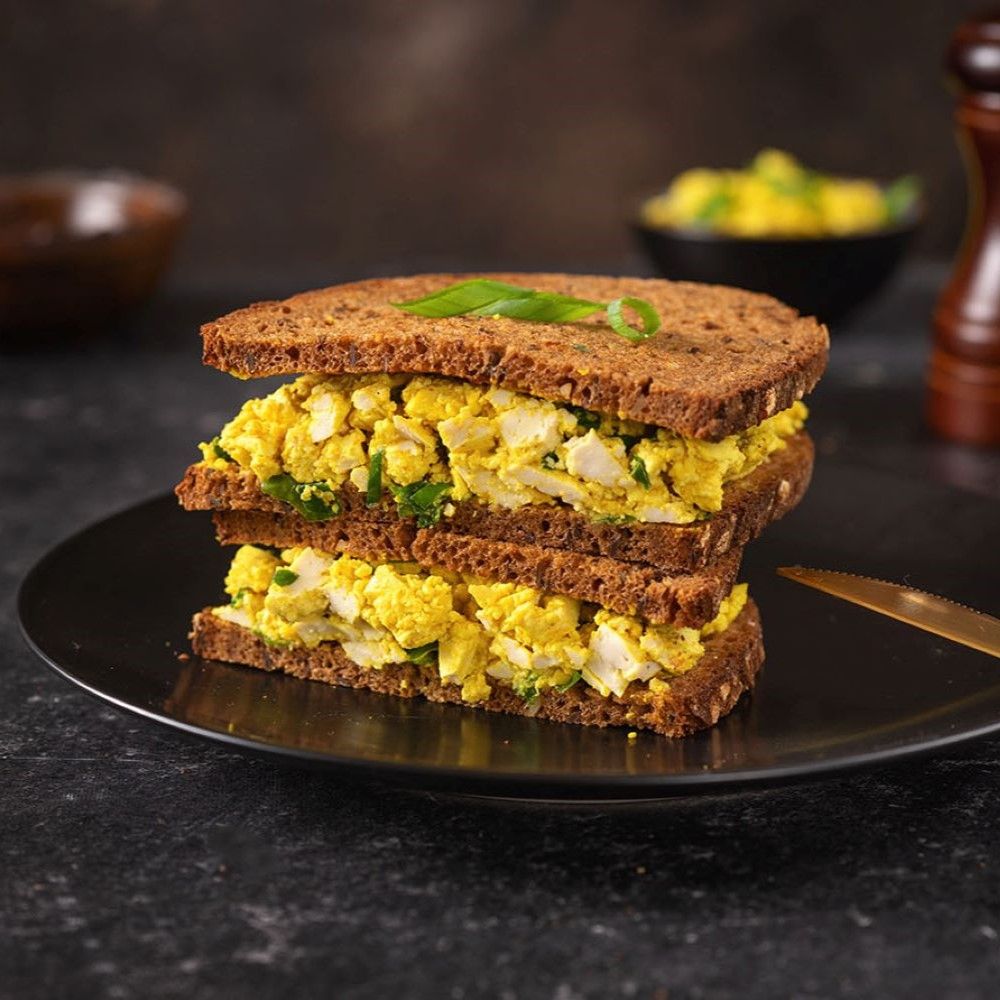 Sandwich mit gelber Füllung auf schwarzem Teller. Schnittlauch. Dunkler Hintergrund.
