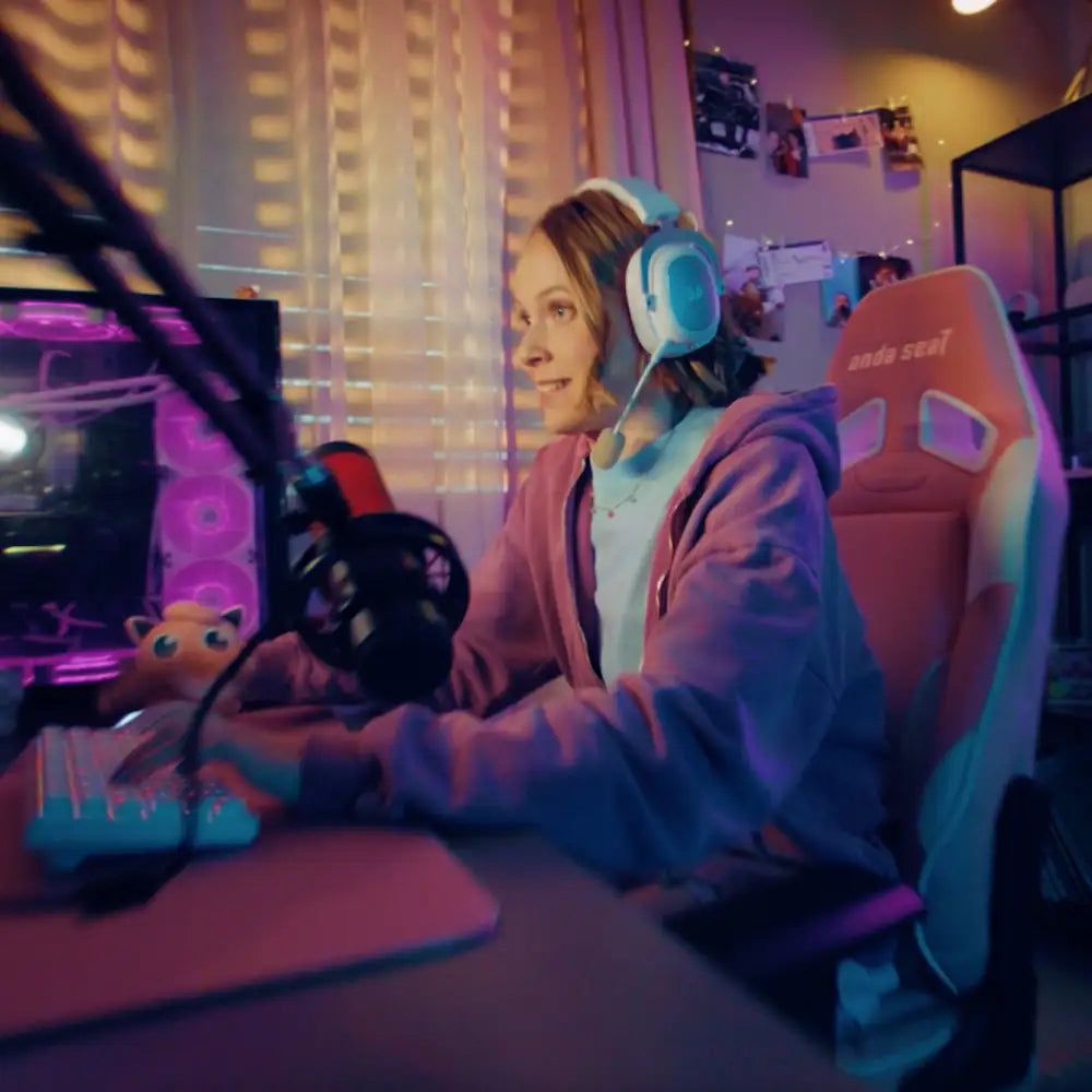 Eine Frau sitzt vor einem Computer und trägt ein Headset. Sie scheint zu spielen oder zu streamen. Im Hintergrund sind Gaming-Accessoires.