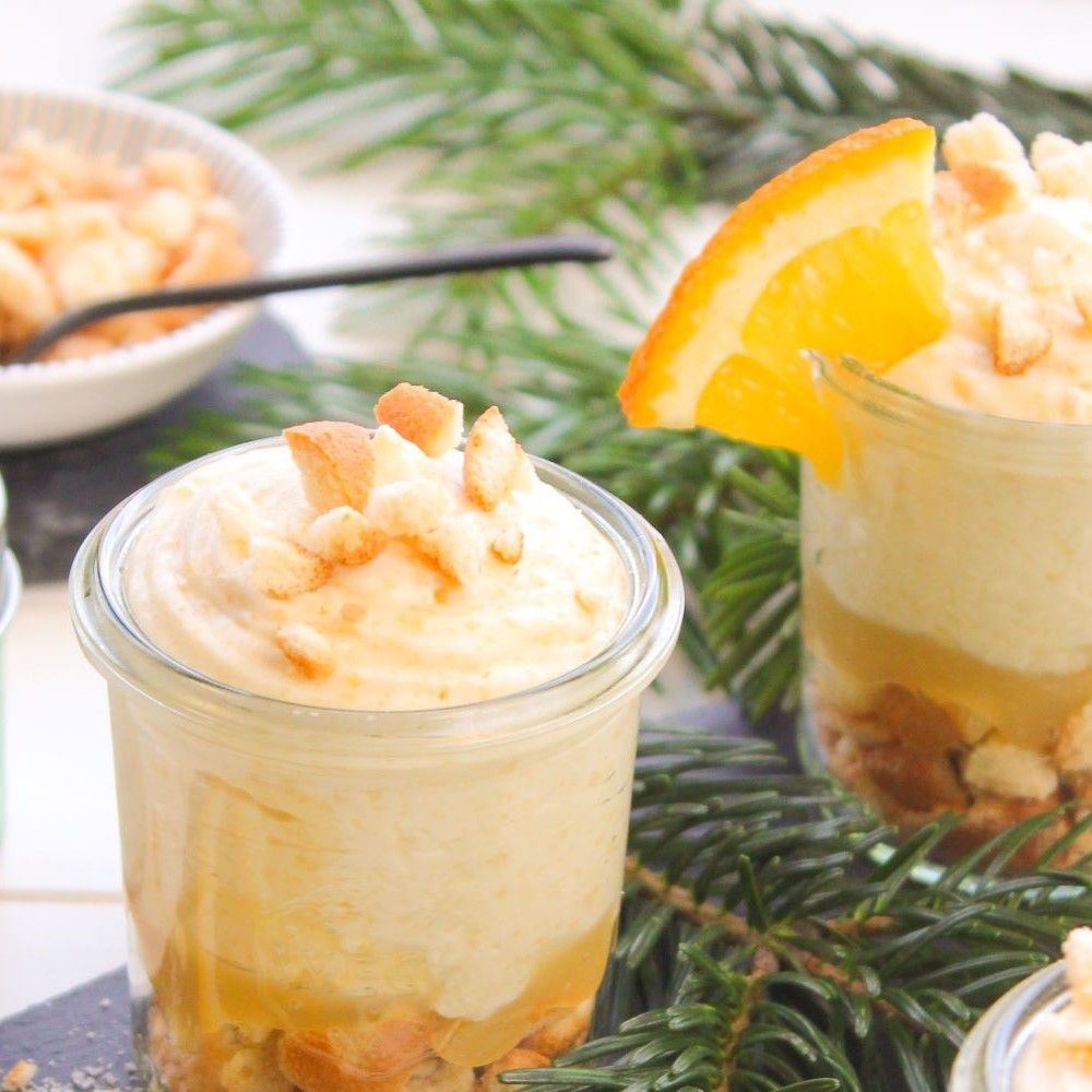 Dessert in Gläsern mit Orangenscheibe. Creme, Kuchenstücke und Orangenscheiben. Grüne Tannenzweige im Hintergrund.
