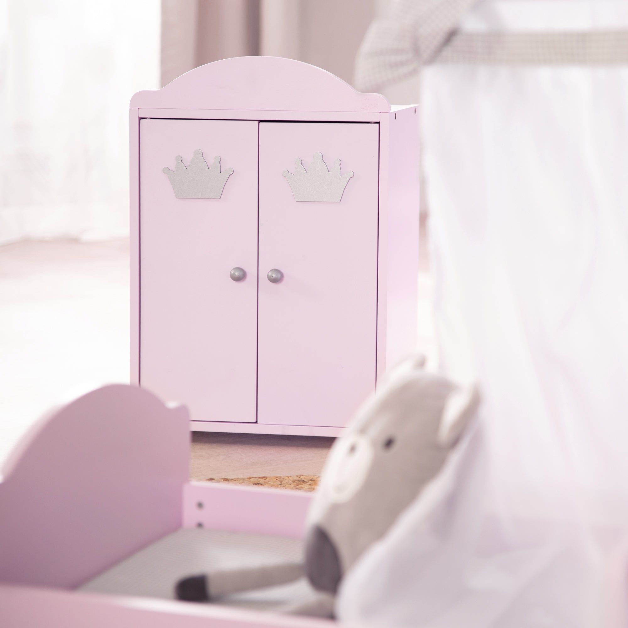 Rosa Puppenschrank mit zwei Türen und silbernen Kronen. Runde Knöpfe. Im Hintergrund ein Bett und ein Stofftier.