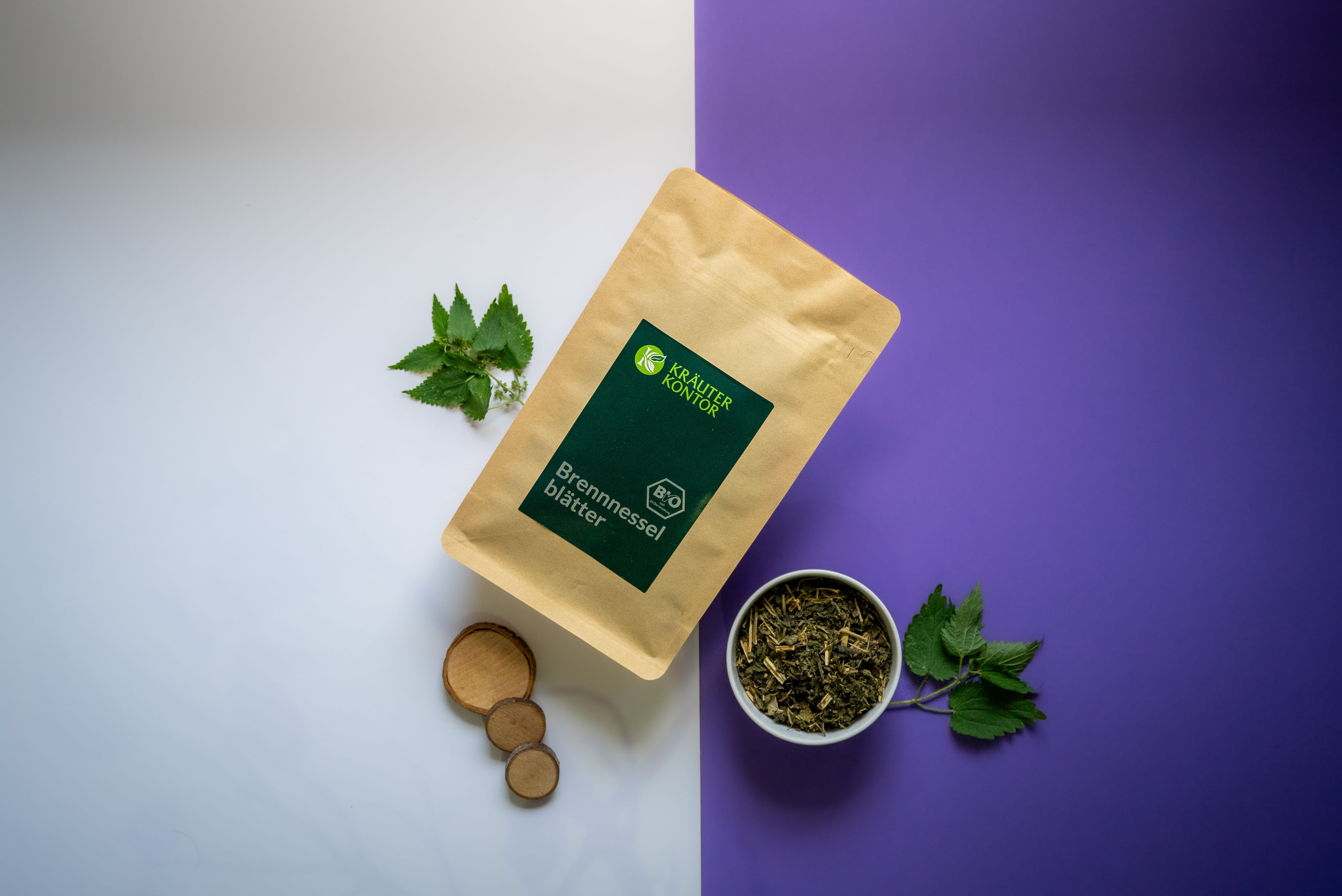 Beutel mit Kräuterkontor Große Brennnesselblätter, grüne Blätter in Schale, Holzscheiben und Brennnesselzweige auf weiß-lila Hintergrund.