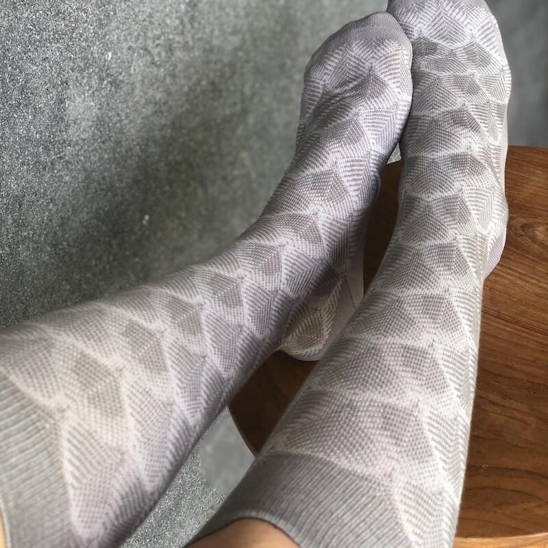 Zwei Beine mit grauen, gemusterten Stützstrümpfen. Geometrisches Muster. Füße auf Holzoberfläche.