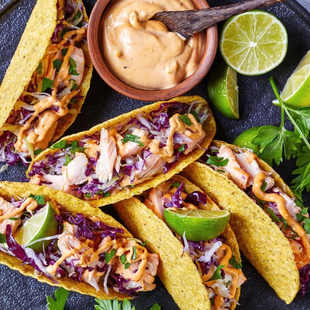 Tacos gefüllt mit Fleisch, Krautsalat und Sauce. Limettenspalten. Auf schwarzem Teller.