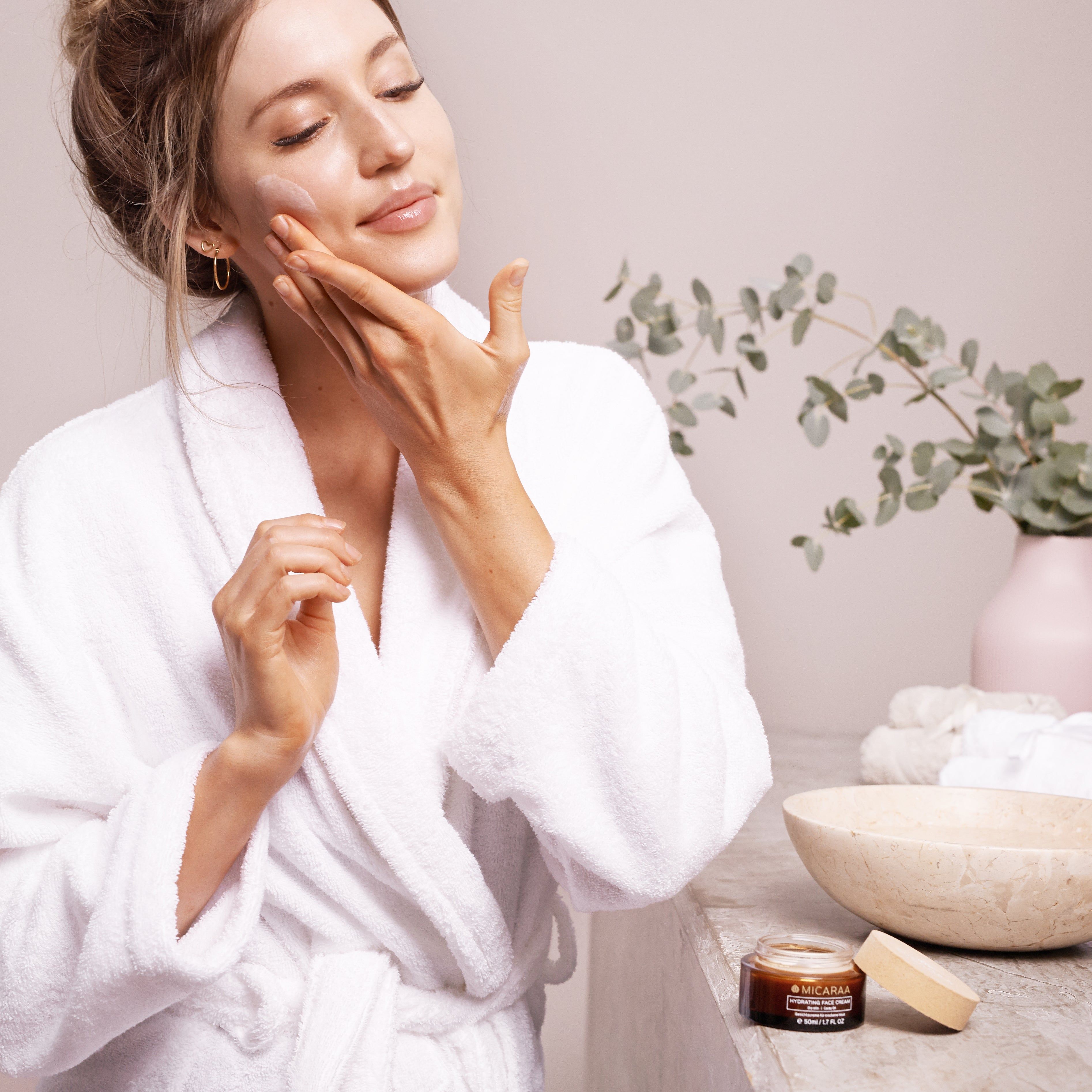 Frau trägt Creme auf. Braune Creme-Dose mit Holzdeckel, Aufschrift: Hydrating Face Cream, Micaraa. Frau im Bademantel.