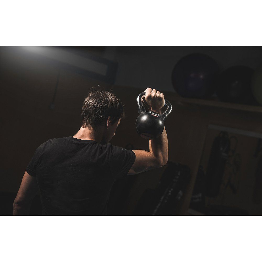 Person hebt eine schwarze Kettlebell mit Chromgriff. Der Griff wird gehalten. Im Hintergrund sind weitere Fitnessgeräte zu sehen.