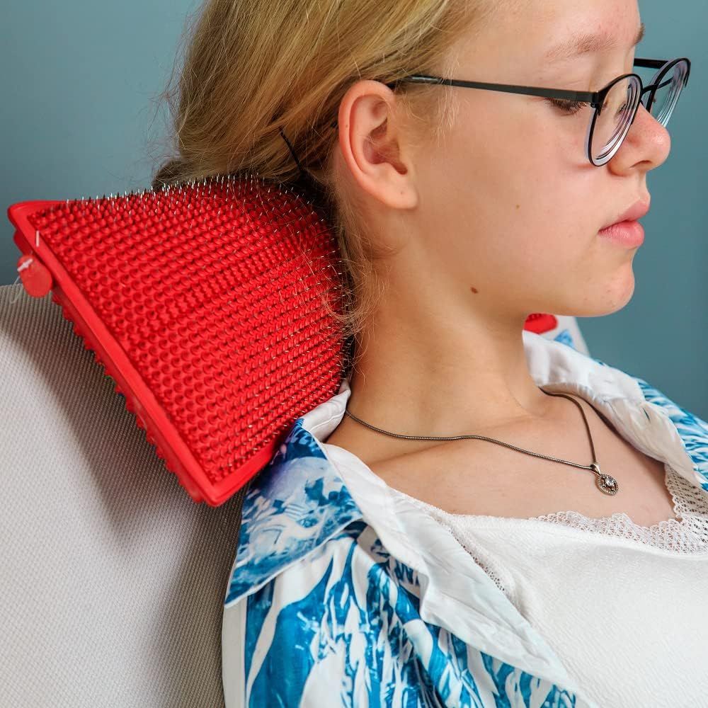 Eine Person mit Brille, die ein rotes, spitzes Kissen am Nacken hält. Die Person sitzt auf einem Stuhl.