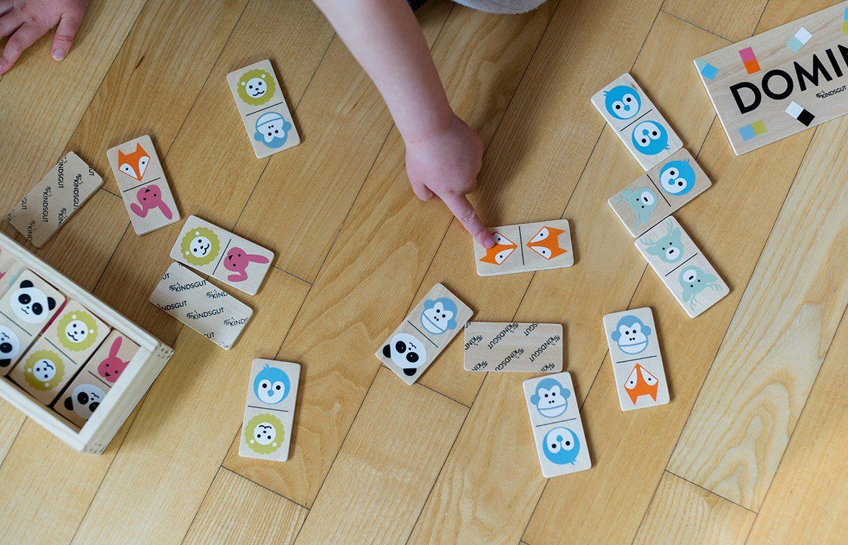 Kind spielt mit Dominosteinen auf Holzboden. Die Steine zeigen Tiergesichter. Eine Holzkiste ist im Bild.
