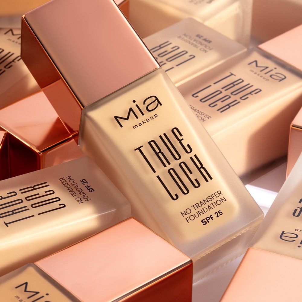 Diverse boccette di fondotinta Mia Makeup True Lock, parzialmente coperte. Le boccette sono in vetro con tappi color oro rosa. Sul davanti sono presenti le scritte "TRUE LOCK", "NO TRANSFER FOUNDATION" e "SPF 25".
