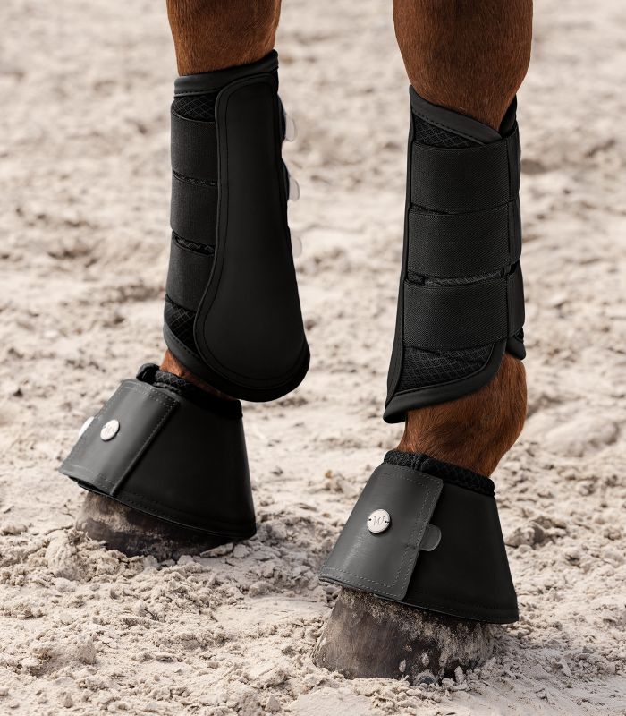 Pferd mit schwarzer Beinbekleidung und Hufglocken. Hufglocken mit Logo. Steht auf Sandboden.