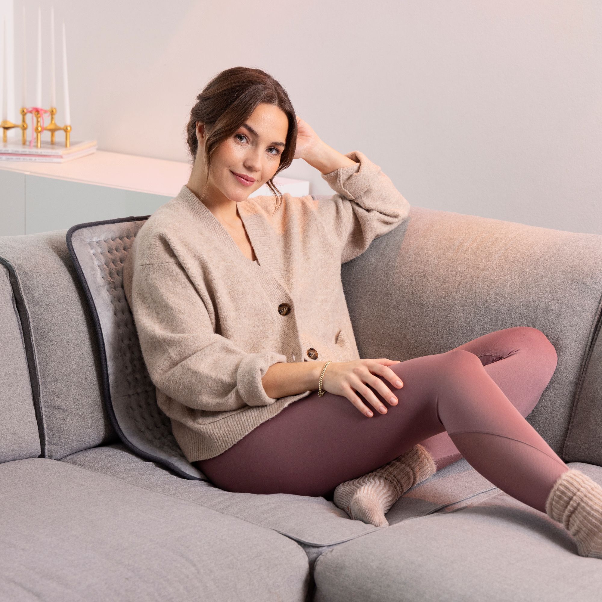 Frau sitzt auf Sofa, Heizkissen unter ihr. Graues Kissen mit schwarzem Rand. Frau trägt Strickjacke und Leggings.