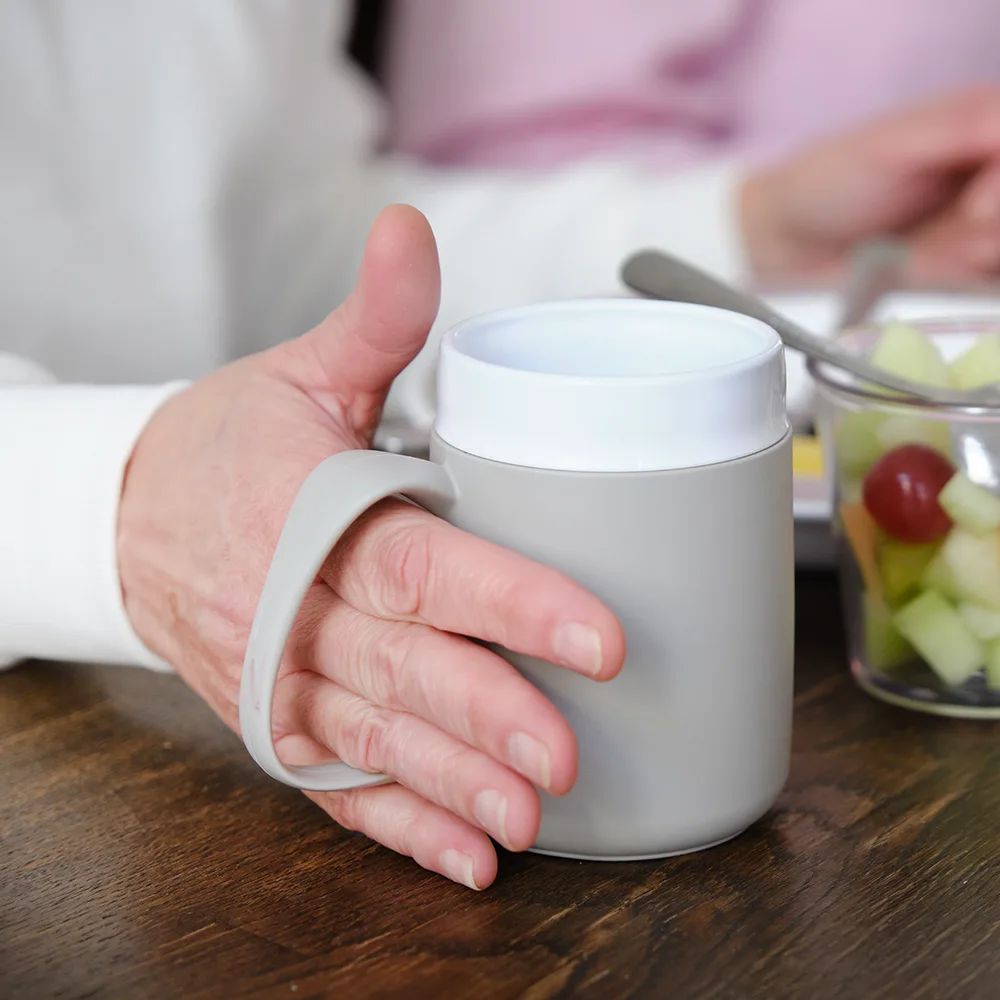 Person hält einen grauen Becher mit großem Henkel. Auf einem Tisch, neben einem Glas mit Obst.