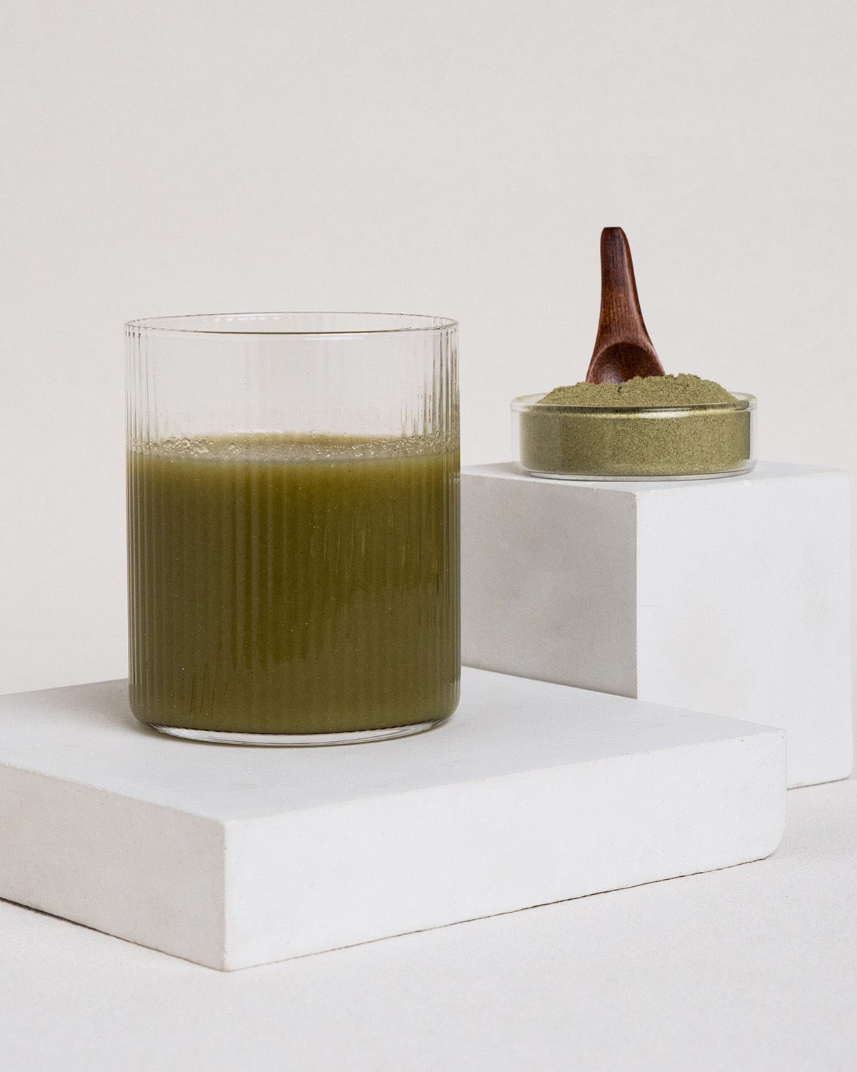 Glas mit grünem Getränk, daneben Schale mit grünem Pulver und Holzlöffel. Auf weißen Blöcken platziert.