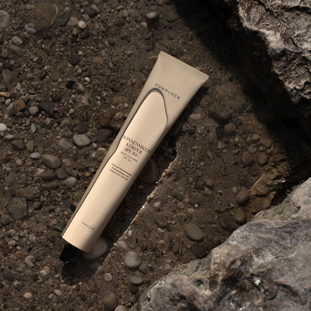 Tube SONNENMILCH KÖRPER SPF 50+ liegt auf Stein. Beige Tube mit schwarzem Verschluss. JUNGLÜCK Schriftzug.