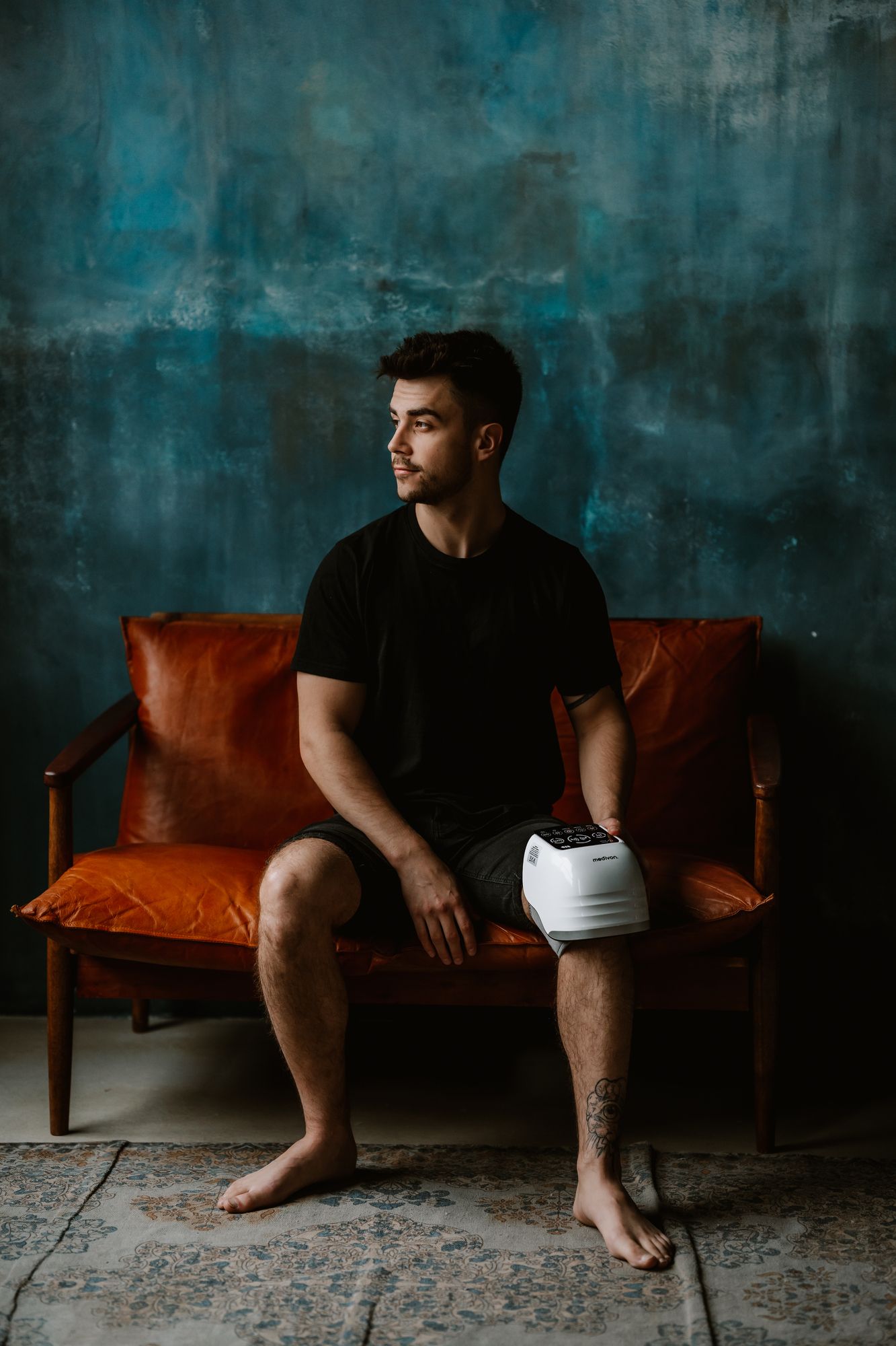 Mann sitzt auf einer Couch und hält ein weißes Knie Massagegerät. Das Gerät ist am Knie platziert.