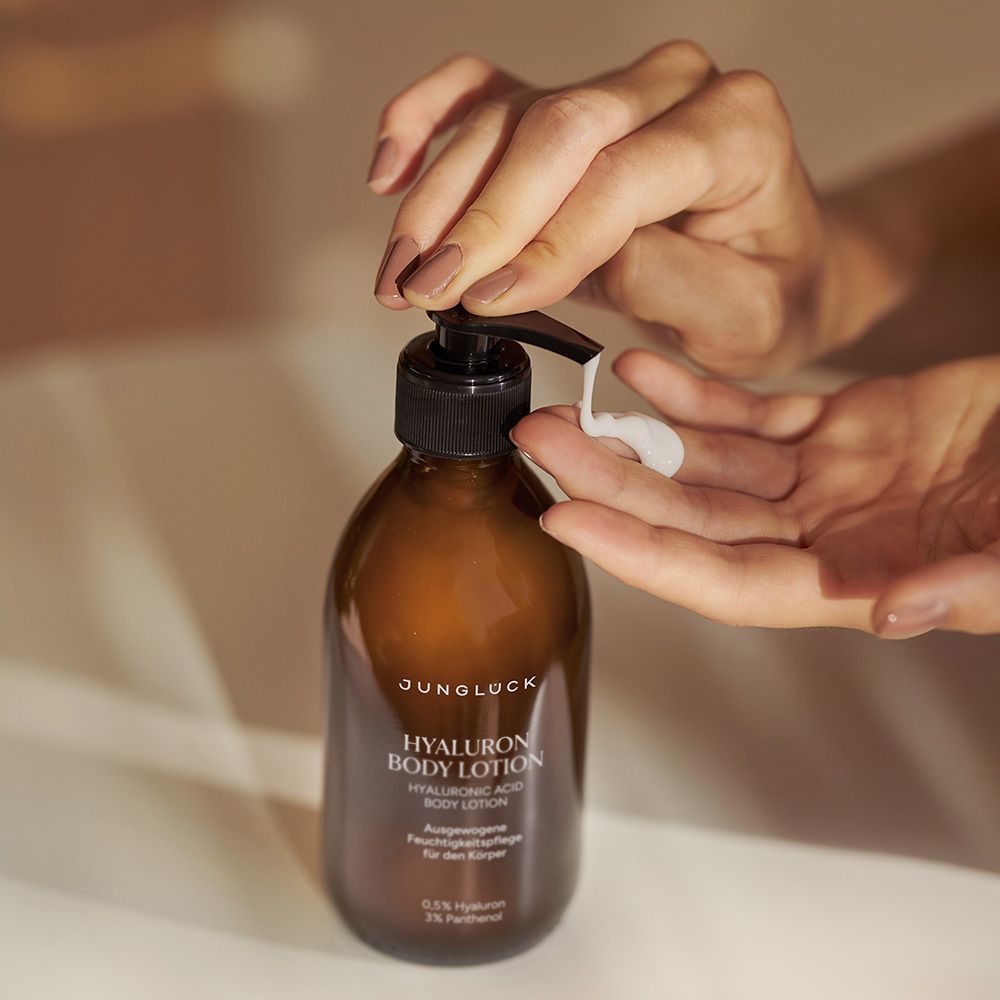 Hände drücken Lotion aus einer braunen Flasche mit schwarzem Pumpkopf. Aufschrift: JUNGLÜCK, Hyaluron Body Lotion. Creme wird auf die Handfläche gegeben.