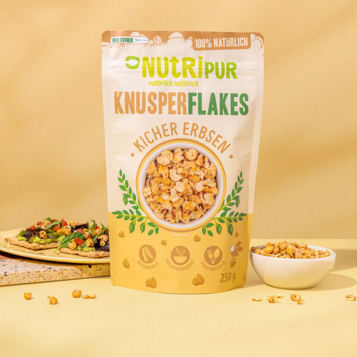 Packung Knusperflakes Kichererbsen, Marke NutriPur. Aufschrift: 100% natürlich. Auf dem Beutel ist eine Schüssel mit Flakes abgebildet. Daneben eine Schüssel mit Flakes.