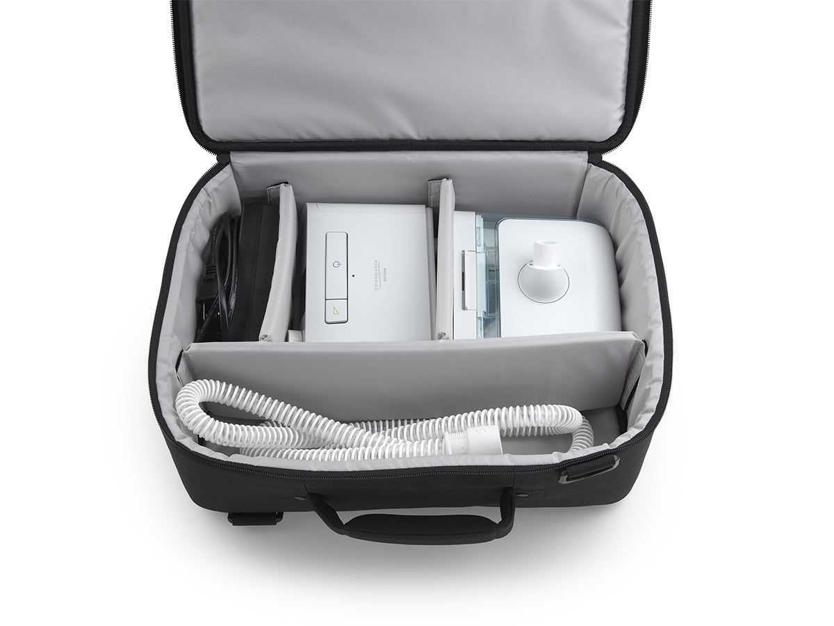 Schwarze Reisetasche, geöffnet, mit weißen Therapiegeräten und Schläuchen im Inneren, aufgeteilt in Fächer.