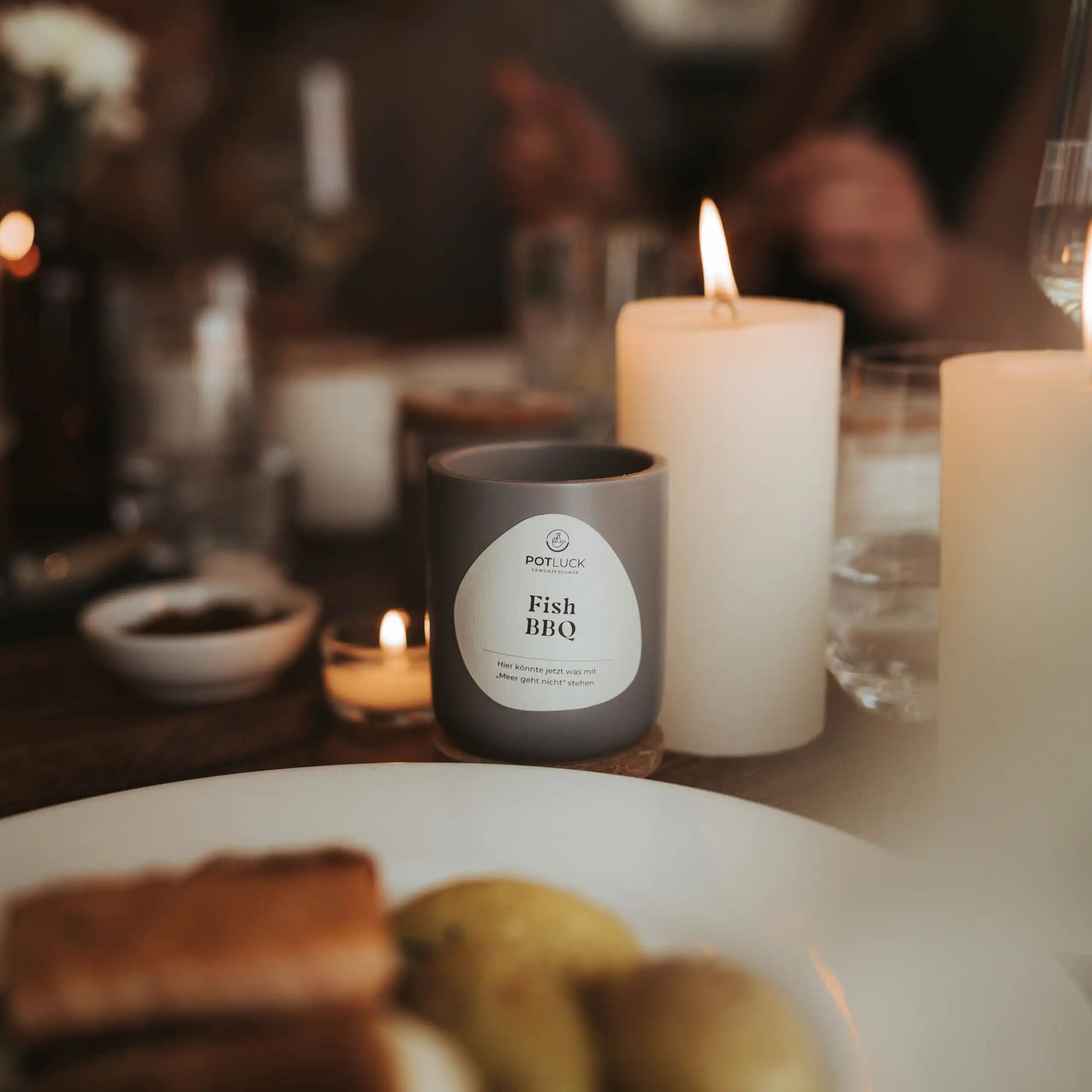 Graue Dose mit Aufschrift POTLUCK Fish BBQ auf einem Tisch, umgeben von Kerzen und Essen.