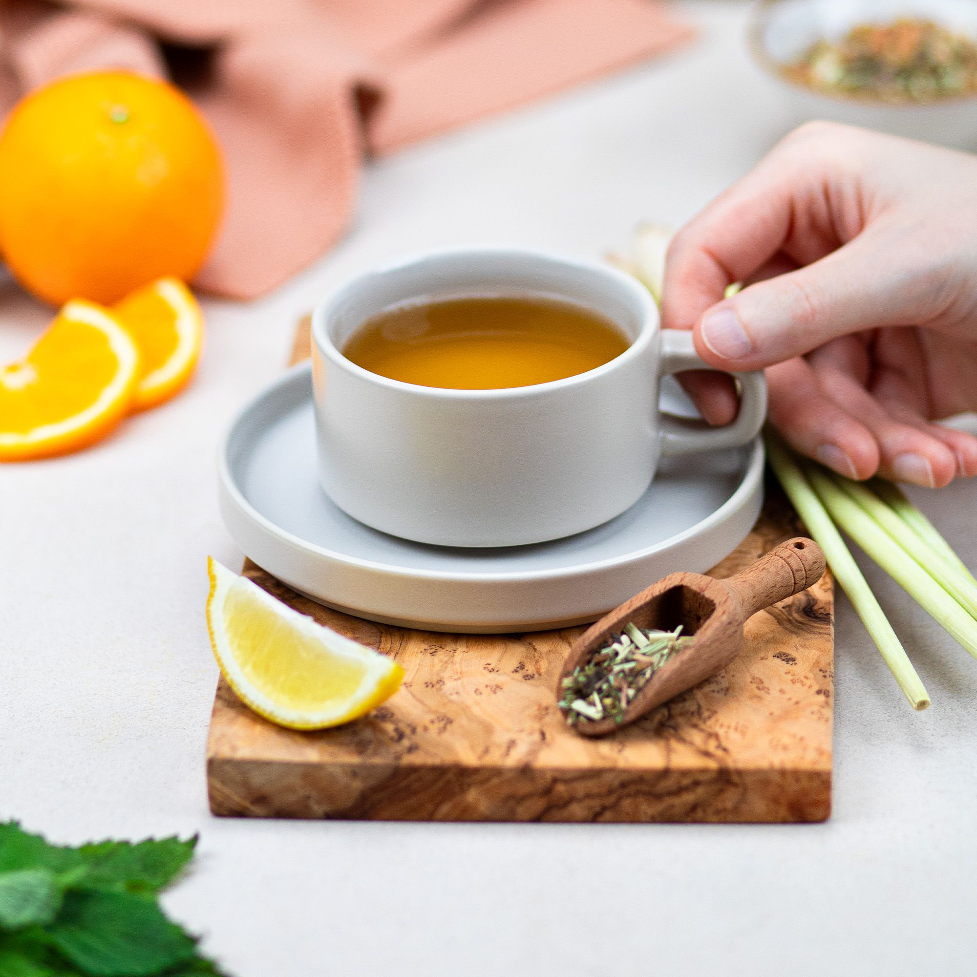 Tasse Tee auf Untertasse, Holzbrett mit Zitrone, Orange, Zitronengras und Holzlöffel mit Tee. Hand hält Tasse.