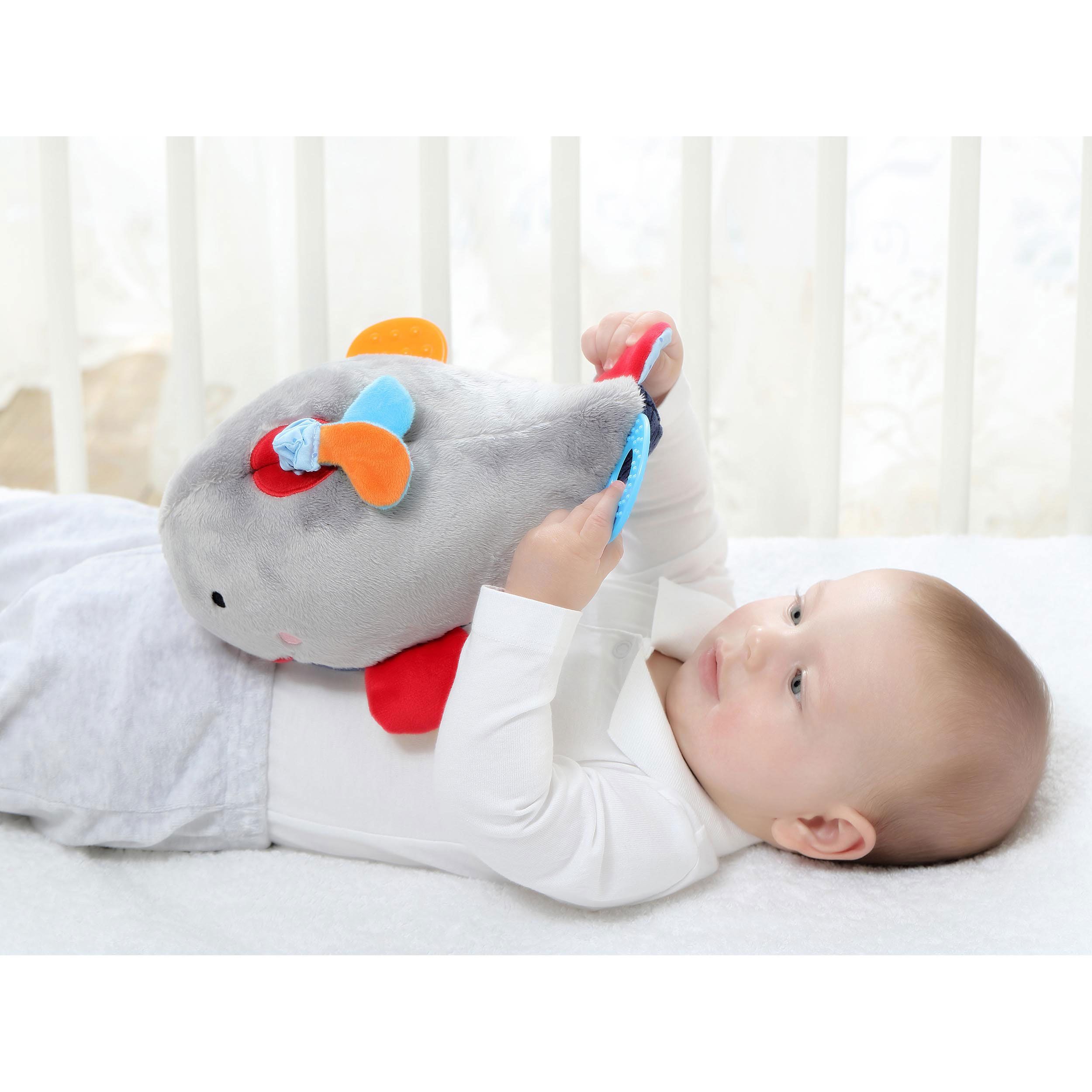 Baby liegt mit grauer Wal-Spielfigur. Rote, blaue und orangefarbene Elemente. Kind hält Ring.