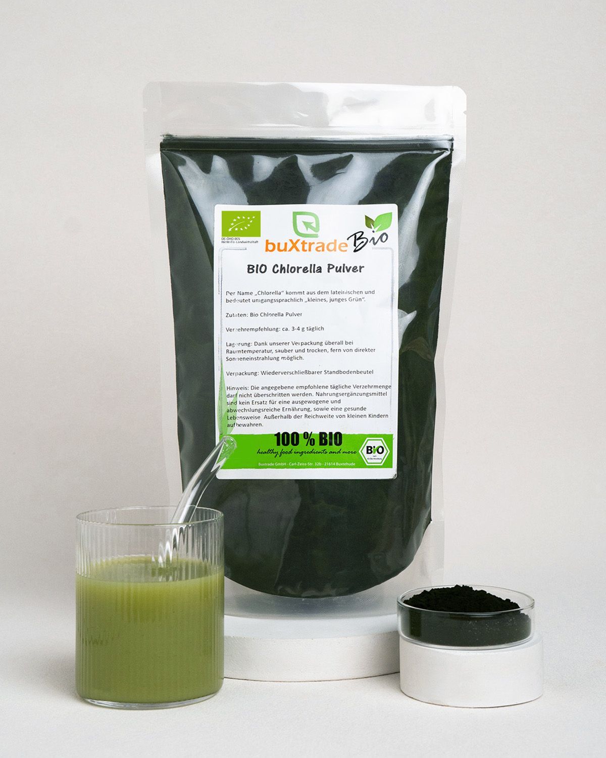 Buxtrade Bio Chlorella Pulver in einem Standbodenbeutel. Daneben ein Glas mit grünem Getränk und eine kleine Schale mit Pulver.