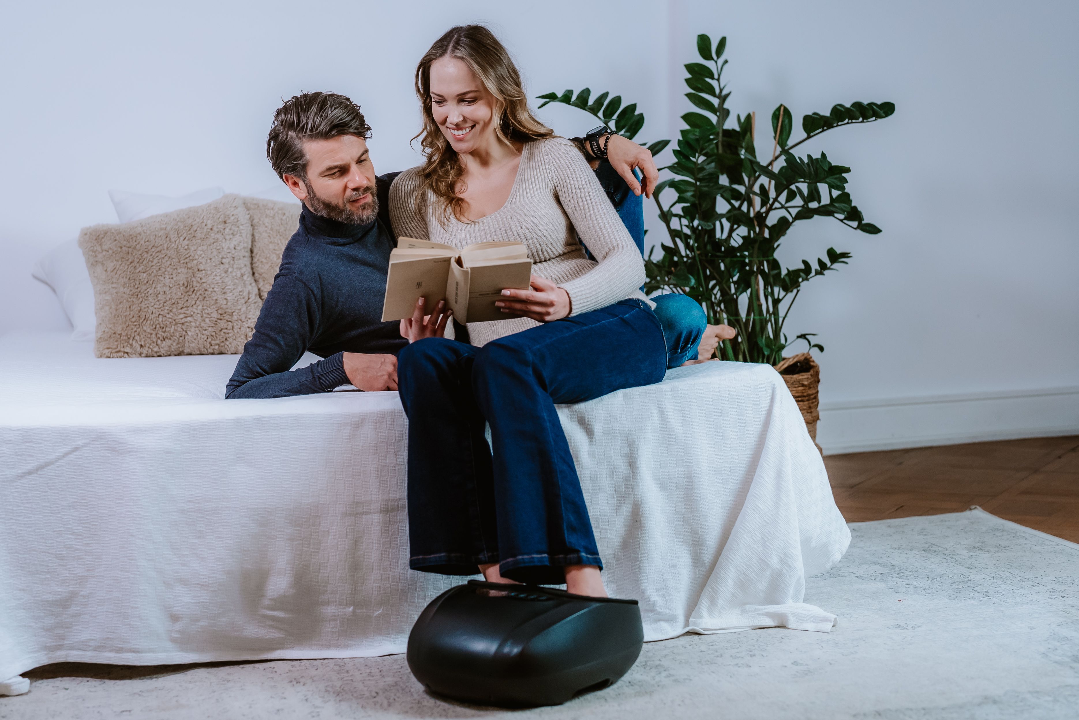 Paar sitzt auf Bett, Frau liest Buch, Füße auf schwarzem Fußmassagegerät.