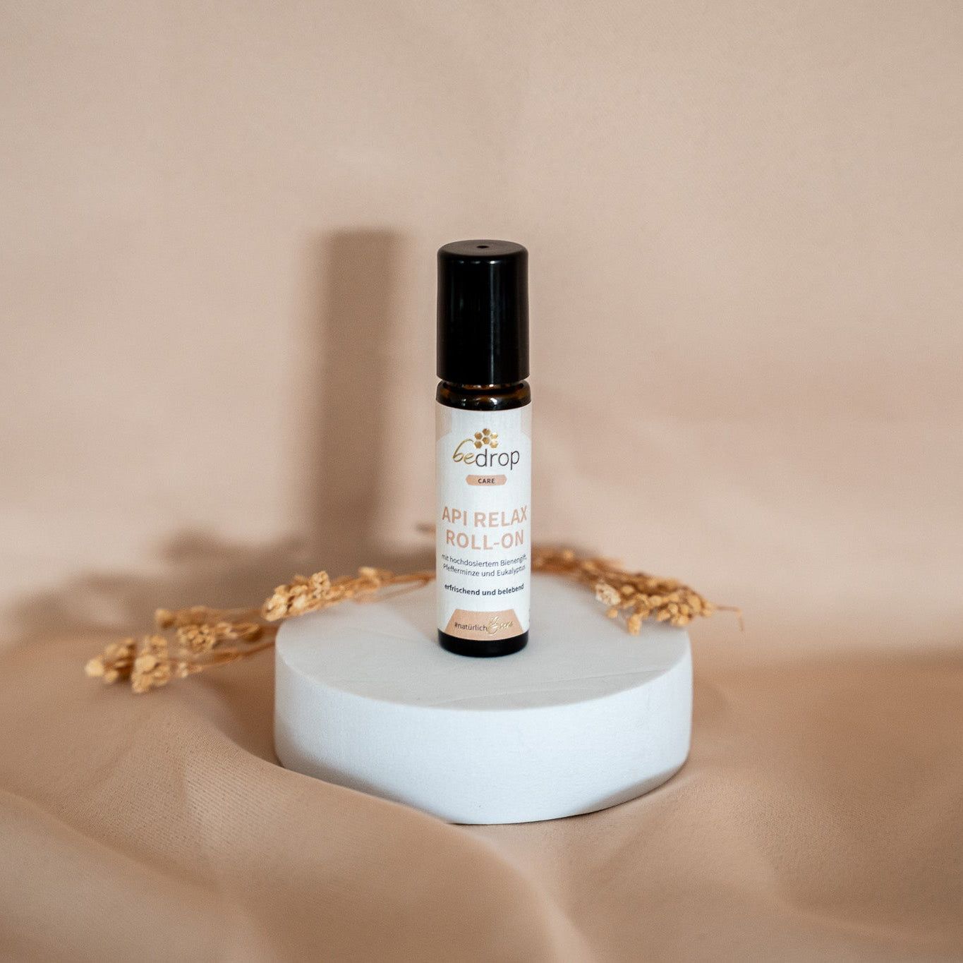 Braune Glasflasche mit schwarzem Deckel auf weißem Podest. Aufschrift: bedrop API RELAX ROLL-ON. Dekoration mit Getreide.