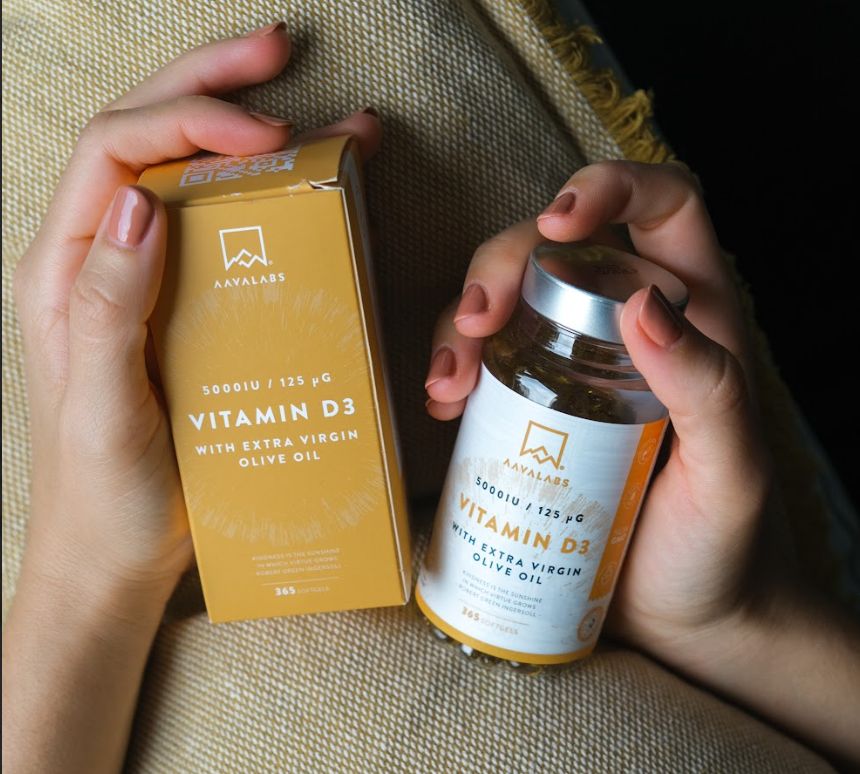 Hände halten eine Schachtel und ein Kapselglas. Aufschrift: Vitamin D3, 5000 IE, mit nativem Olivenöl. Gelber Hintergrund.