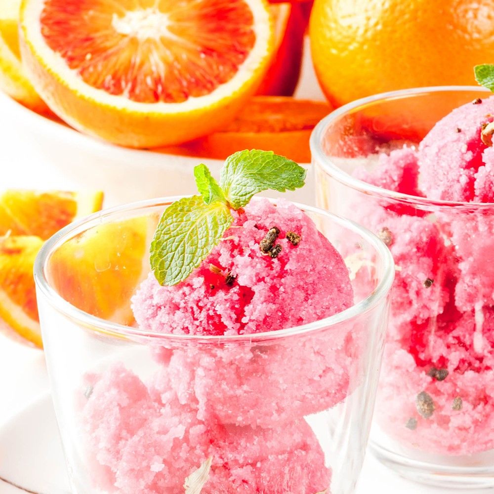 Zwei Gläser mit rosa Eiscreme, garniert mit Minze. Orangen und Blutorangen im Hintergrund.