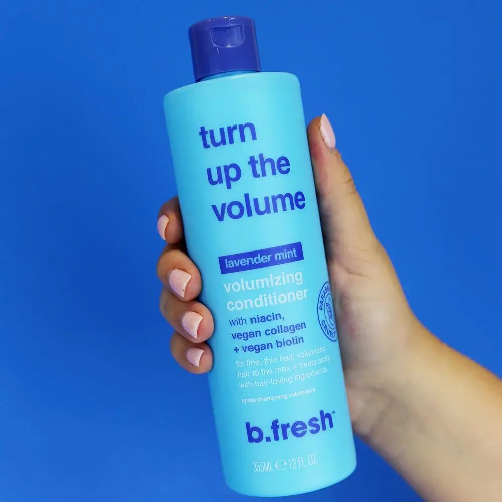 Hellblaue Flasche mit blauem Deckel, gehalten von einer Hand. Aufschrift: Turn Up the Volume, Volumizing Conditioner. Enthält Niacin, veganes Kollagen, Biotin.