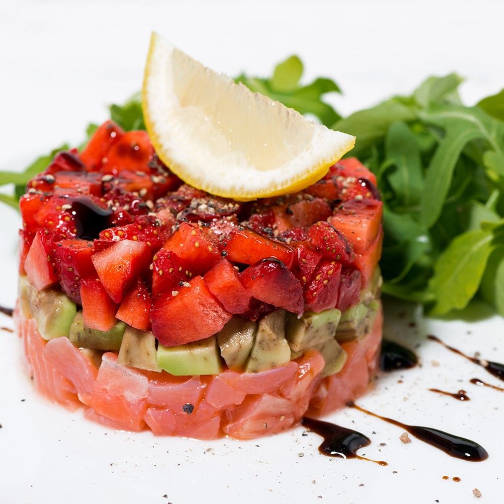 Türmchen aus Lachs, Avocado und Erdbeeren, mit Zitrone und Rucola. Balsamico-Glasur auf weißem Teller.