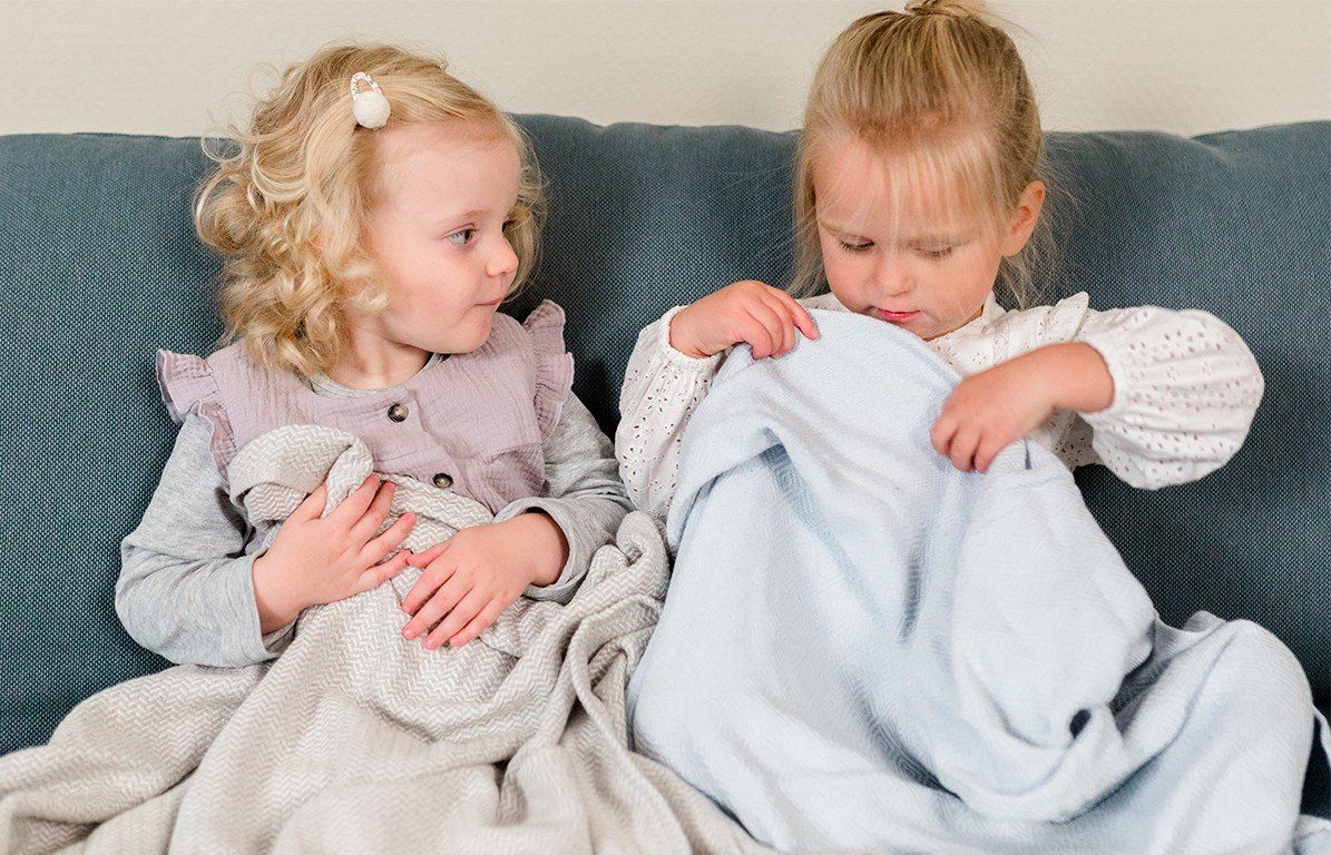 Zwei Kleinkinder sitzen auf einem Sofa. Ein Kind hält eine hellblaue Decke.