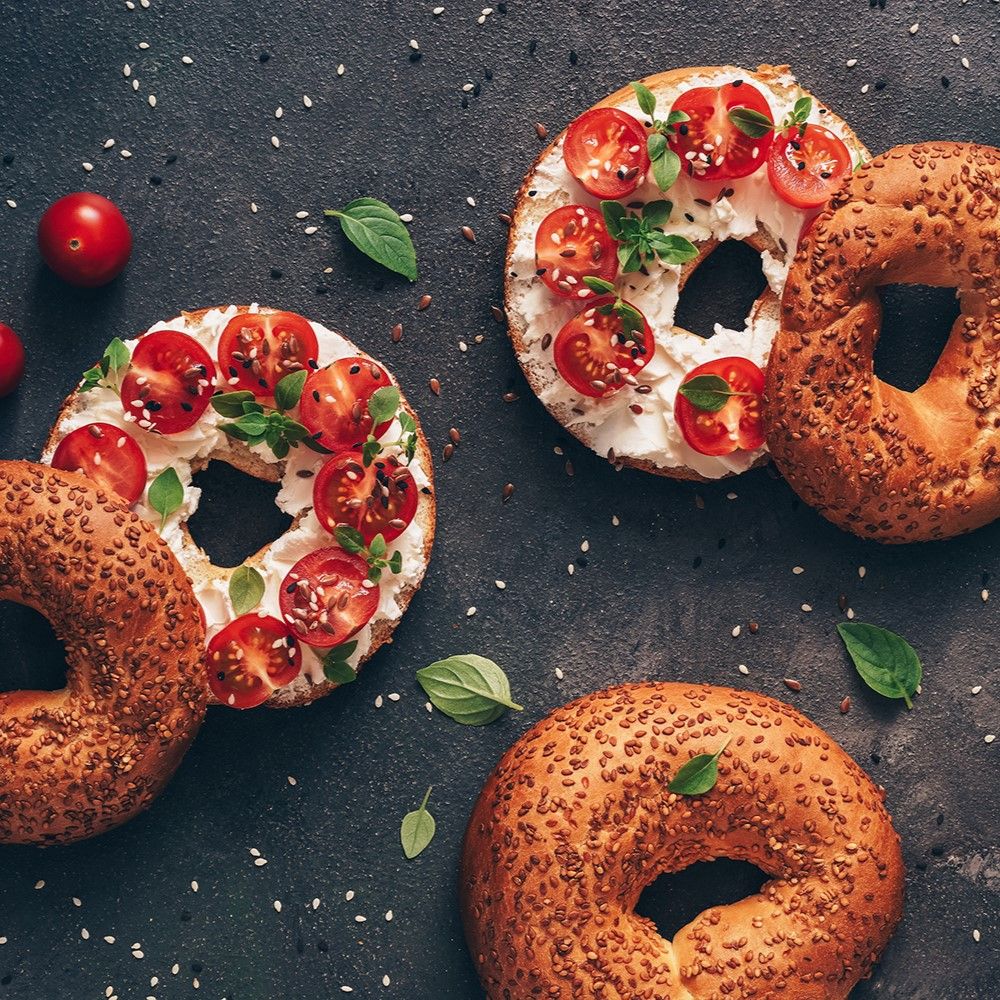 Bagels mit Frischkäse, Tomaten und Basilikum. Einige Bagels sind mit Sesam bestreut. Draufsicht auf dunklem Untergrund.