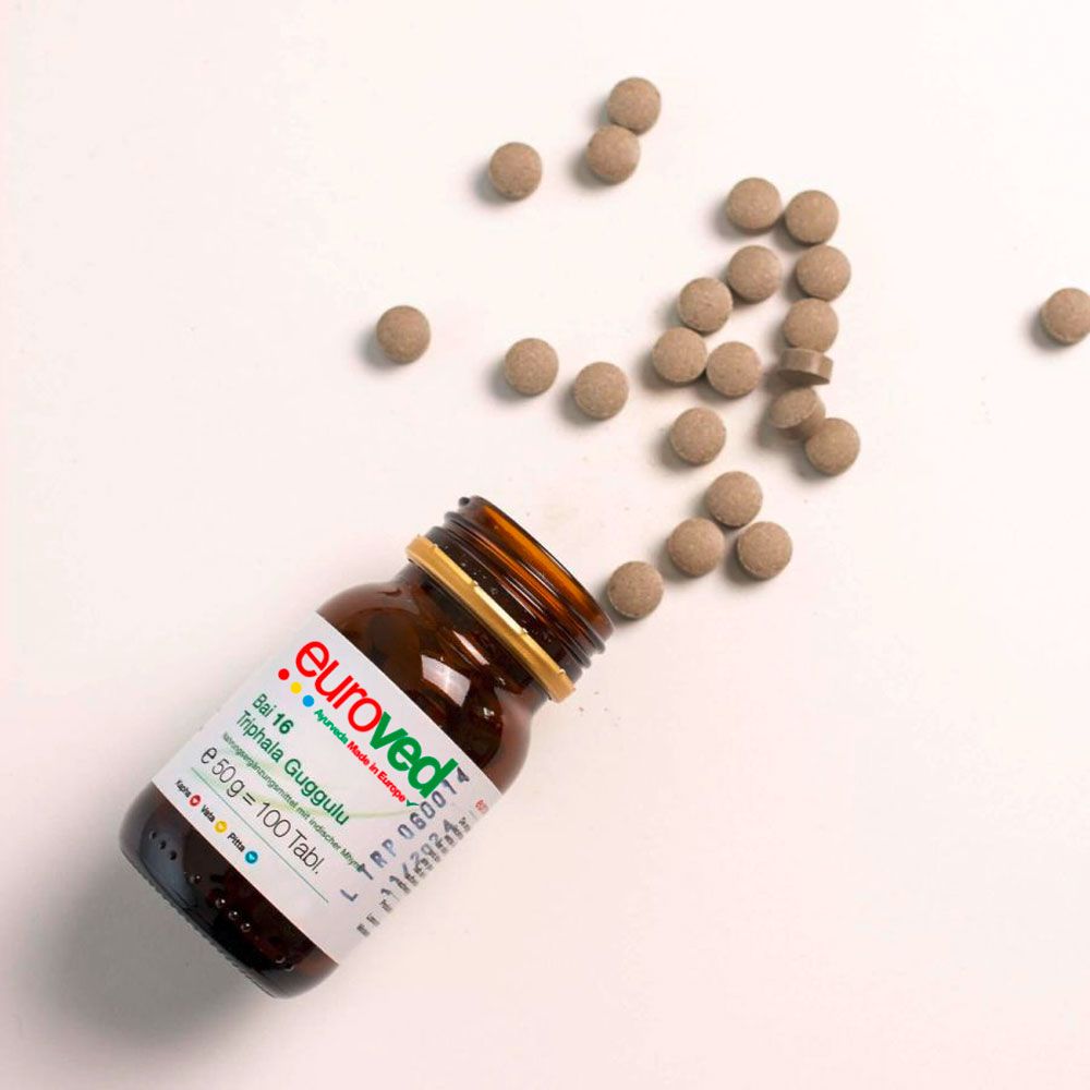 Braune Glasflasche, teilweise geöffnet, mit Tabletten daneben. Etikett mit Produktnamen und Informationen. "euroved" und "Bai 16 Triphala Guggulu" sichtbar.