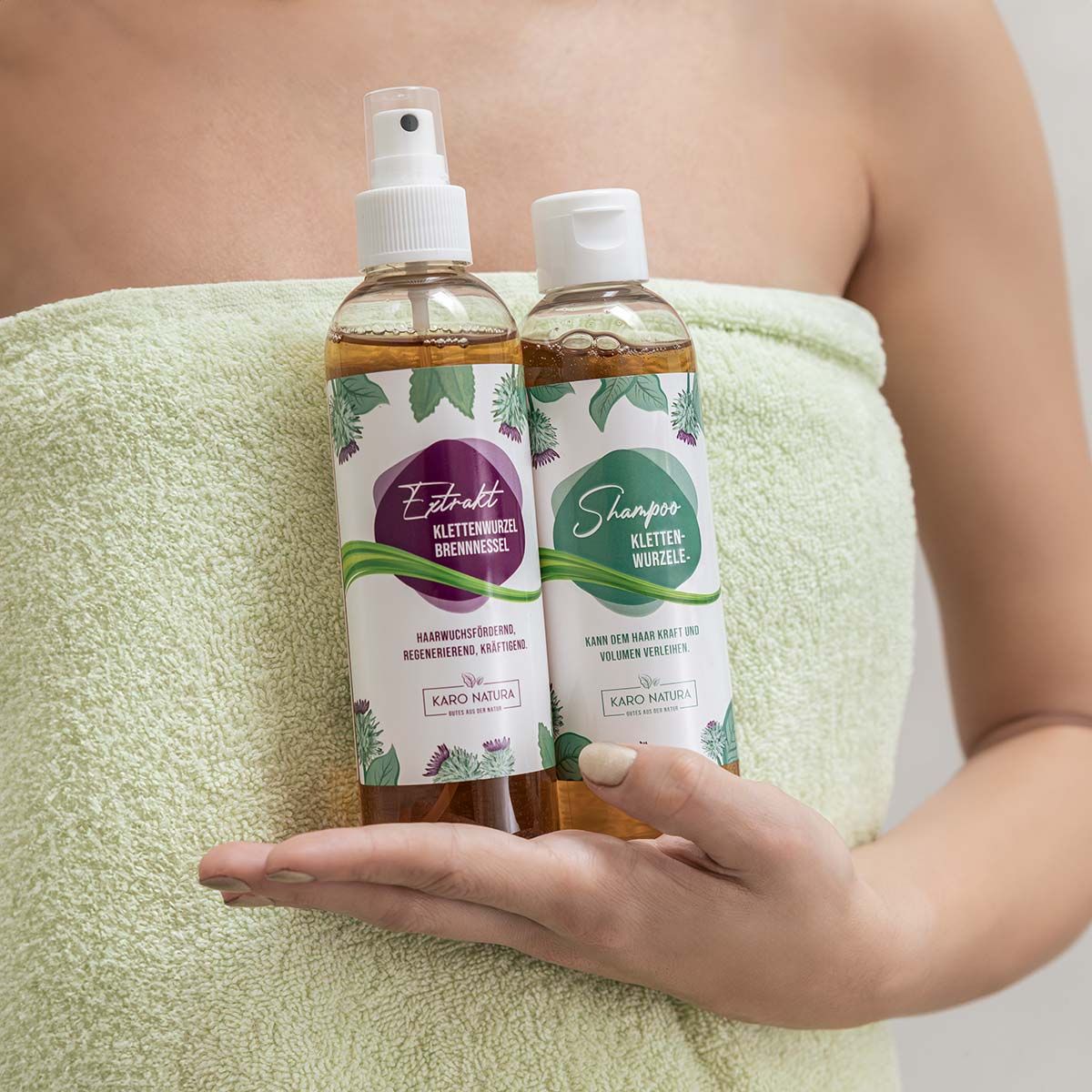Zwei Flaschen, eine mit Sprühkopf, gehalten von einer Person. Etiketten: Extrakt und Shampoo, Karo Natura.