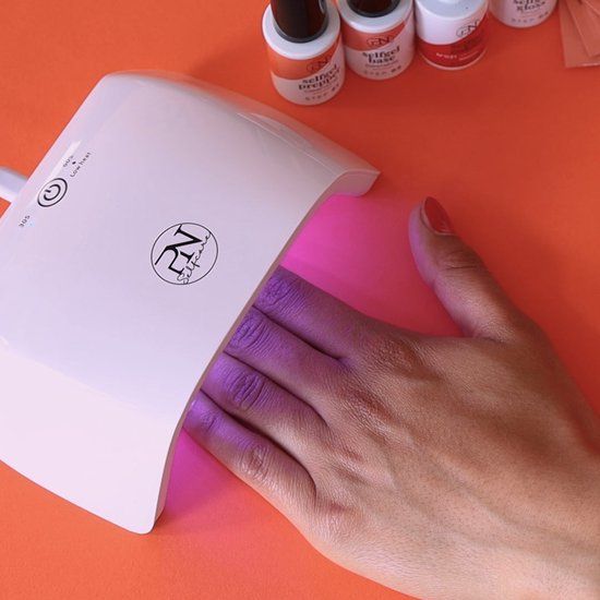 Hand in UV-Nagellampe. Lampe weiß, mit PN-Logo. Nägel mit rosa Gel. Hintergrund orange.